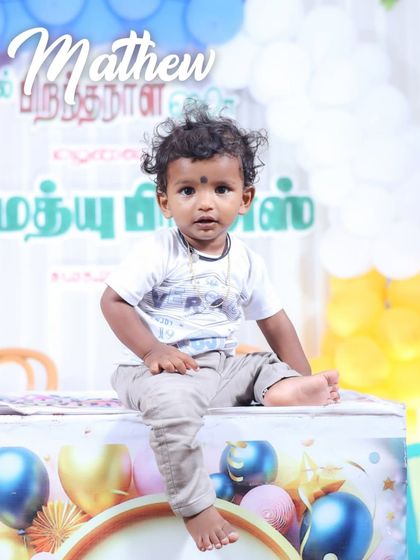 A portrait of baby Matthew at his birthday celebration. Seated in front of a colorful balloon backdrop with his name in Tamil, this shot captures the festive atmosphere of the event.
