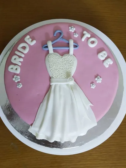A sweet and charming cake perfect for a bridal shower. This design features a fondant wedding dress with pearl details on a hanger, set against a lovely pink background with "Bride to Be" lettering.