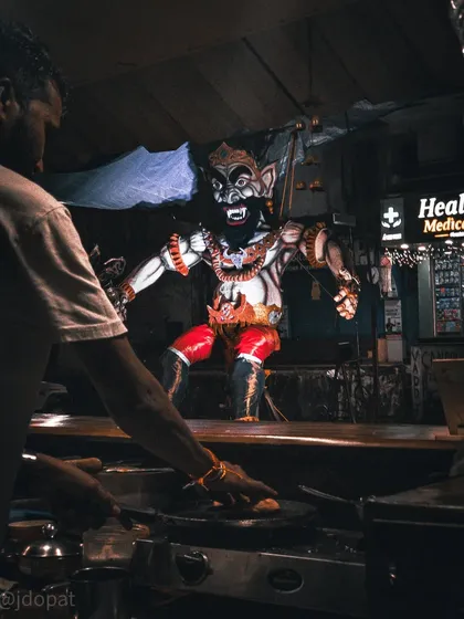 A street food vendor works at his stall, with the imposing figure of a Narakasura effigy looming in the background.
