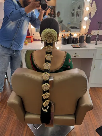 A behind-the-scenes shot of Ragavi's traditional braid being photographed. The hairstyle features a full poola jada with intricate gold ornaments, a masterpiece of bridal hairstyling.