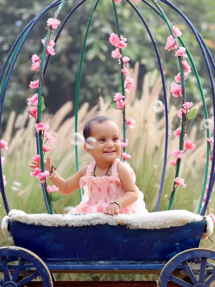 A smiling baby girl sitting in a whimsical, flower-adorned carriage prop in a beautiful garden setting.