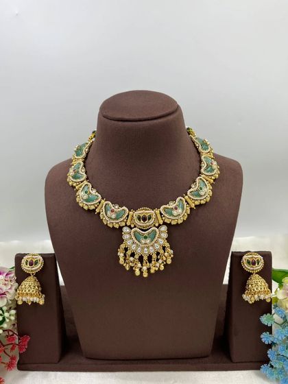 The Tishyaa Gold & Gems necklace. This piece showcases beautiful green Meenakari work in a crescent shape with a detailed pendant and matching jhumkis, all in a rich antique gold finish.