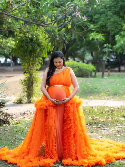 A full-length shot of a mom-to-be in a vibrant orange gown, standing in a park and lovingly holding her baby bump.