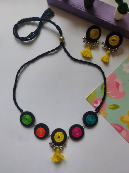 A playful and colorful handmade necklace set. This design features thread-wrapped circles in multiple colors on a black cord, with matching earrings that have bright yellow tassels.