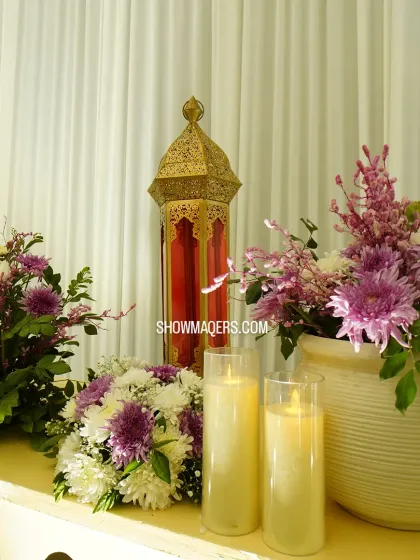 A close up of a floral arrangement featuring purple chrysanthemums and white flowers, set beside a traditional lantern and candles.