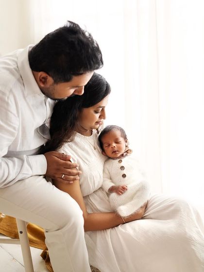 A quiet, candid moment as a family of three sits together, admiring their new baby. The soft window light adds a beautiful, gentle quality to the photo.