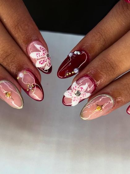 An elegant and artistic set combining a deep maroon base with sheer accent nails. The look is elevated with hand-painted white orchids and sparkling gold details.