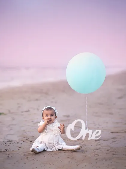 A whimsical first birthday portrait on the beach, with a single balloon adding a pop of color against the pastel sky.