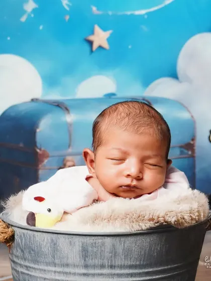 A sleeping newborn in a metal bucket, with a dreamy blue background featuring clouds and stars.