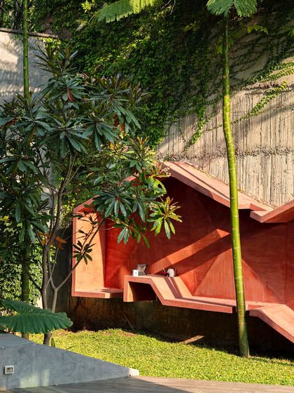 The red sculptural bench seen from a distance, nestled in a corner of the lawn against a concrete and vine-covered wall. Its bold form and color make it a striking focal point in the garden.