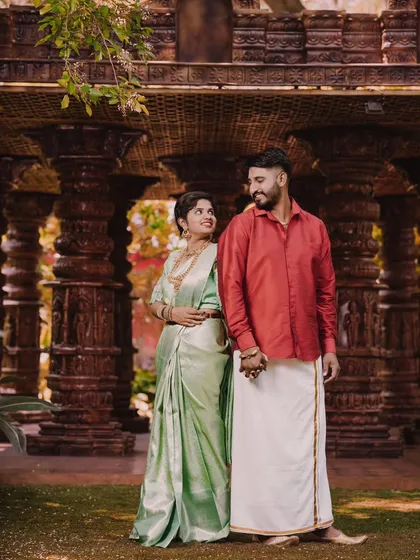 A beautiful pre-wedding shot of a couple in traditional South Indian attire, standing amidst intricately carved wooden pillars that evoke a temple's architecture.