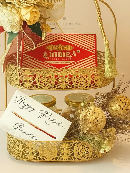 A detailed view of the two-tiered Rakhi caddy, showing the gourmet dry fruits and Bombay Sweet Shop mithai. The golden baubles and dried flowers add a festive, celebratory touch.