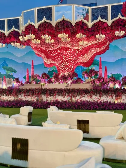 A wide shot of the surreal stage and lounge area. The modern white furniture provides a stark, beautiful contrast to the vibrant colors of the floral decor and the green lawn.