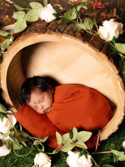 A close-up of a newborn wrapped in a rust-colored swaddle, nestled inside a hollow log prop and adorned with white roses.