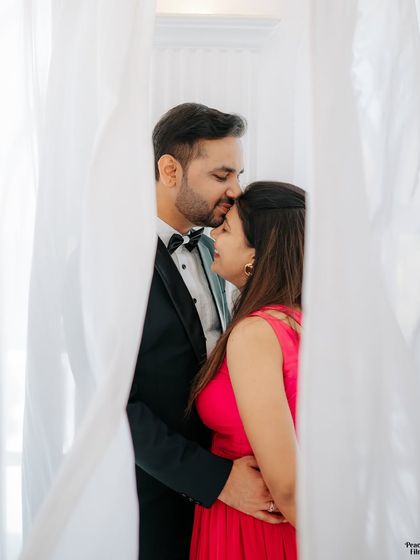 A tender pre-wedding portrait framed by soft white curtains. The groom's gentle kiss on the bride's forehead is a perfect expression of love and affection.