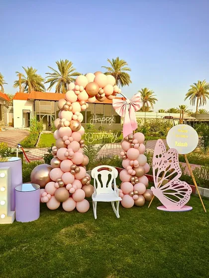 A beautiful butterfly-themed first birthday in a garden. This setup features a pink and gold balloon arch, a large butterfly cutout, and a neon 'Happy Birthday' sign.