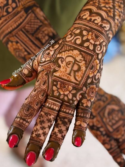 A close-up of a bridal henna design featuring the initial 'R' inside a square frame, surrounded by detailed floral work.
