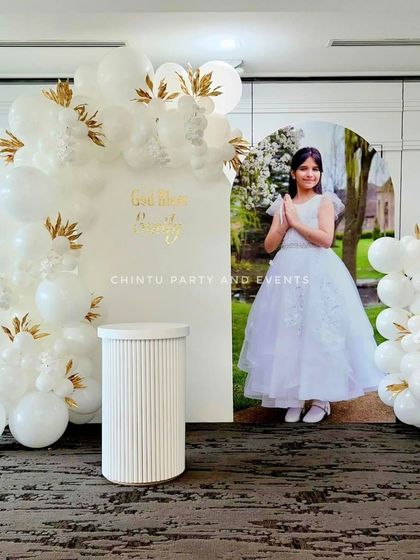 An all-white elegant Holy Communion decoration. The setup includes a personalized backdrop with a photo of the child and a white balloon arch with gold leaf accents.
