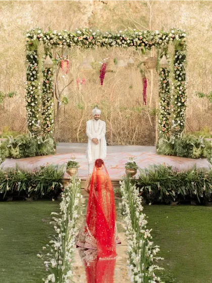 The bride's walk to the mandap is one of the most emotional moments of a wedding. Here, she walks a path lined with flowers towards the groom, who waits under a beautiful floral arch set against a rustic, natural rock wall.