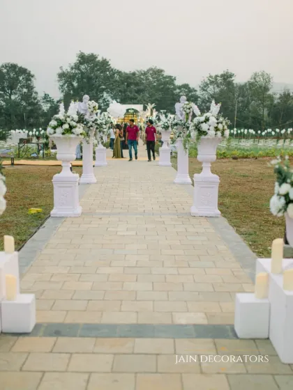 A clean and elegant walkway for an outdoor ceremony. The path is lined with white floral urns and candle-lit pedestals, creating a simple and stately aisle for the couple's entrance.