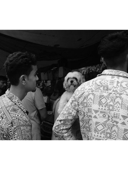 A black and white shot from Sarojini Market where a small dog, held by its owner, seems to be observing the crowd. The matching patterns on the two men's shirts add a unique, almost symmetrical element to the scene.
