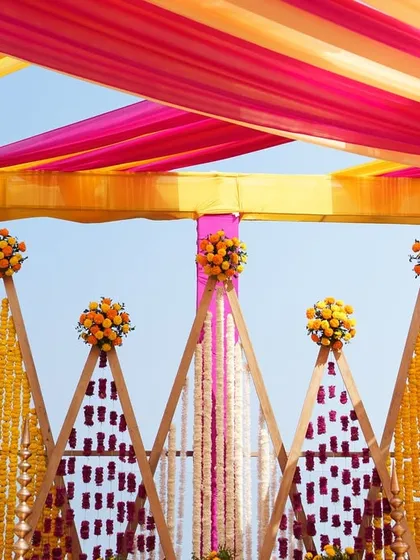 A close-up of the Haldi stage backdrop, featuring triangular frames adorned with strings of yellow, purple, and white flowers.