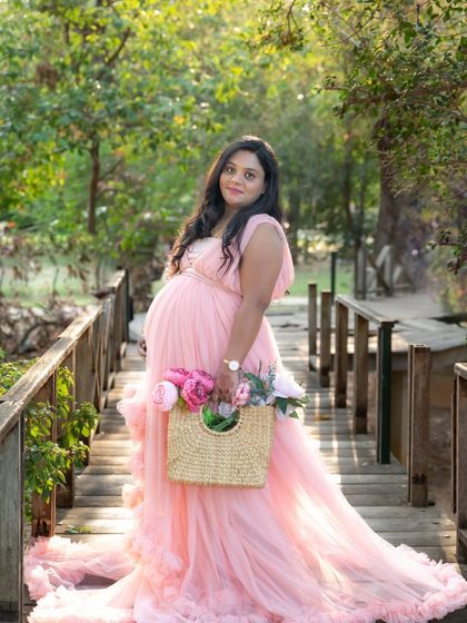 A charming outdoor portrait of a mom-to-be in a pink gown, walking across a wooden bridge while holding a basket of flowers.
