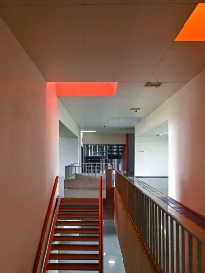 Colored light filters through skylights into a stairwell, painting the walls with hues of red and orange. This use of colored glass transforms the quality of the light, adding an element of surprise and delight to the space.