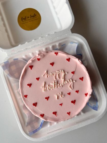 A minimalist pink bento cake with a simple "happy birthday M." message and scattered red hearts. It's a perfect example of a sweet and understated design.