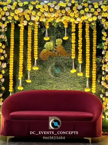 A vibrant baby shower stage with a backdrop of green turf and yellow marigold garlands. The red sofa adds a pop of color, creating a cheerful and festive setting to celebrate motherhood.