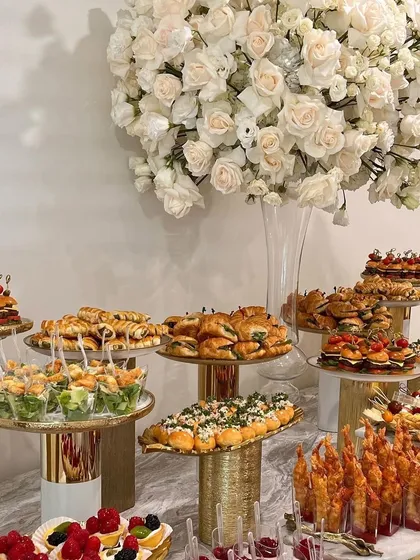 An elegant and formal catering setup. This display features a variety of hors d'oeuvres on tiered platters, complemented by a large white floral arrangement, perfect for a sophisticated reception.