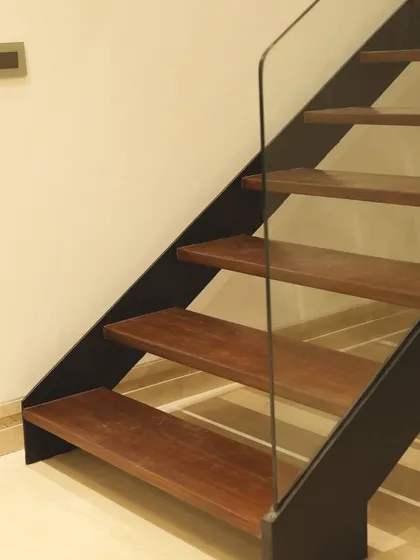 Detail of a minimalist staircase, featuring wooden treads supported by a sleek black metal stringer and a frameless glass railing. This design emphasizes structural honesty and clean lines.