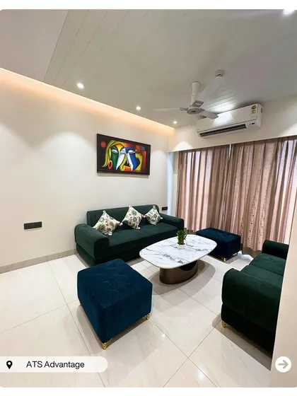 A cozy and contemporary living room setup in a 3BHK at ATS Advantage. The dark green sofa pairs beautifully with blue ottomans and a marble-top coffee table for a balanced, modern look.