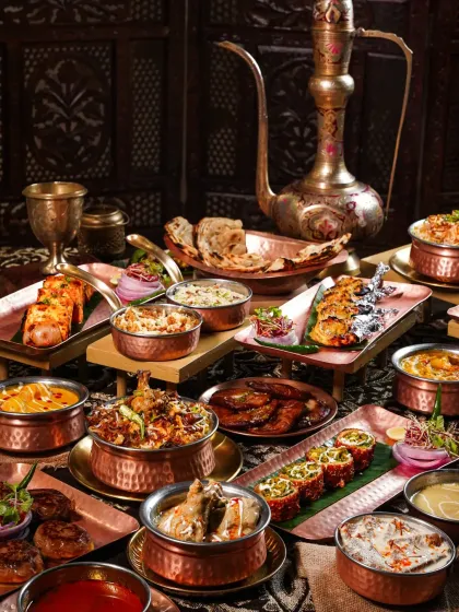 A lavish spread of Awadhi cuisine, featuring biryani, kebabs, and curries in copper and brass vessels. The dark, opulent styling with an antique ewer evokes the royal heritage of Lucknowi food.