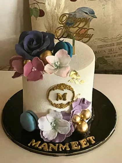 A stylish single-tier cake decorated with a cluster of sugar flowers in shades of purple and pink, macarons, and touches of gold leaf.