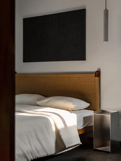 This master bedroom features a custom bed with a handwoven, coffee-colored headboard. The tactile quality of the weaving adds warmth and craft, contrasting with the cool concrete floor and aluminum side table.