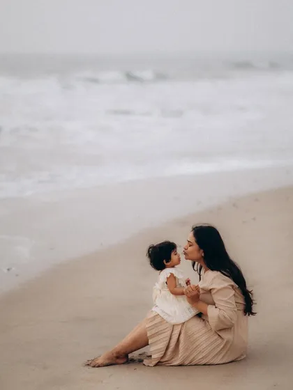 A mother sits on the sand, lifting her baby up for a kiss against the backdrop of the sea. This is a classic, loving pose that feels both timeless and spontaneous in a beach setting.