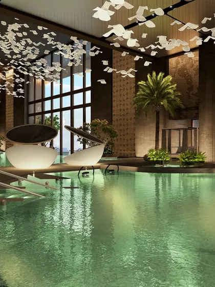 The serene indoor pool at Palais Royale, where poolside leisure is paired with resort ease. The space is framed by wide promenades and palm-lined greenery, with a material palette of honed stone, warm accents, and softly gleaming metal details.