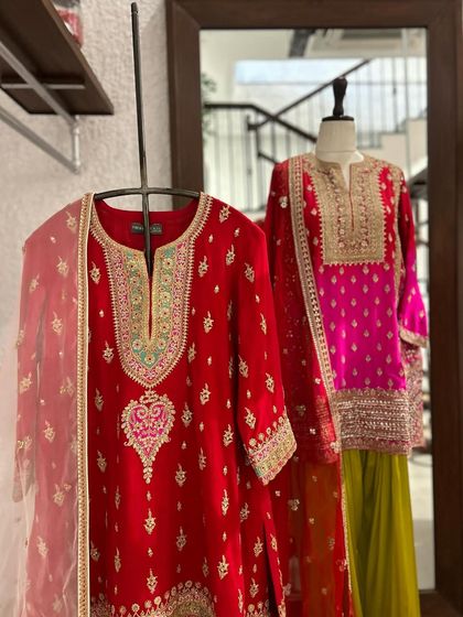 A bold red kurta set with delicate embroidery hangs beside a vibrant pink and lime green sharara set on a mannequin, ready for a festive occasion.