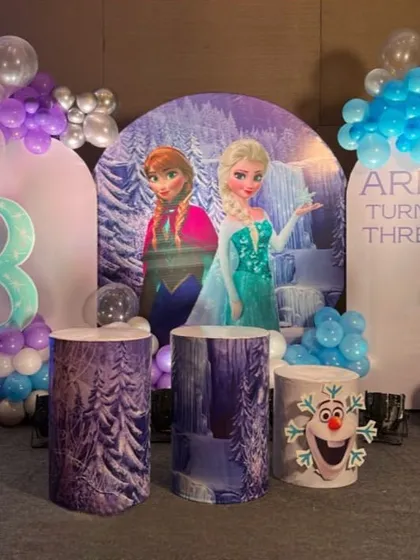 This Frozen-themed birthday decoration features a winter wonderland backdrop with Anna and Elsa. The setup includes themed plinths, including one with Olaf, and a balloon arch in icy blue, purple, and white.