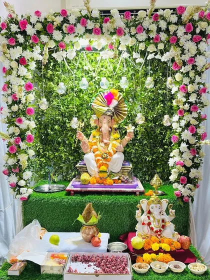 A fresh and green-themed Ganpati decoration for a home celebration. The idol is placed against a lush green wall, framed by an arch of white and pink roses, creating a natural and peaceful setting.