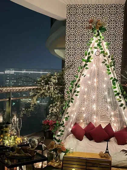 We create magical moments with romantic proposal decorations. This intimate balcony setup features a beautiful teepee canopy illuminated with fairy lights against the city skyline, setting the perfect mood for a 'yes'.
