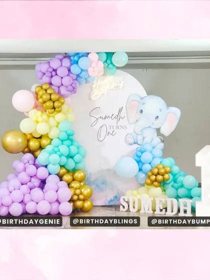 A dreamy pastel elephant theme for a first birthday. The setup features a large balloon arch in purple, yellow, mint, and pink, with a cute baby elephant graphic and a light-up number '1'.