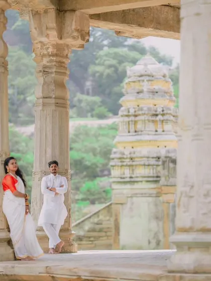A wide shot of a couple posing amidst the stone pillars of a historic temple, with the gopuram visible in the background. This composition emphasizes the grandeur of the location.
