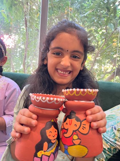 A student proudly shows off the pair of terracotta pots she painted with adorable Lakshmi and Ganesha figures for Diwali.