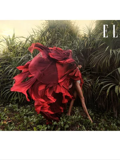 A dramatic, motion-filled shot for an Elle editorial. The model is enveloped in a swirl of vibrant red fabric, becoming one with the wild, natural landscape. This image is pure visual poetry, focused on color, movement, and emotion.