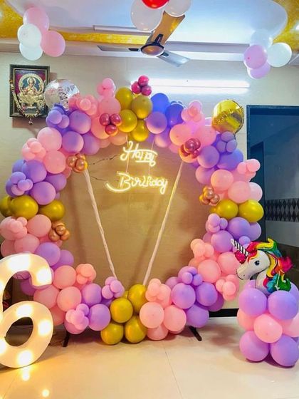 A circular balloon arrangement in pink, purple, and yellow for a 6th birthday, featuring a neon 'Happy Birthday' sign and a small unicorn prop.