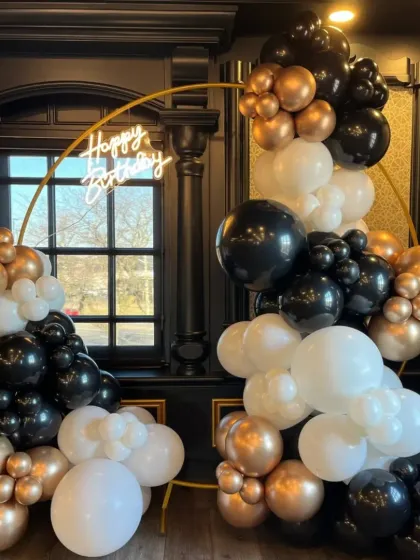 This chic birthday setup features a golden ring arch with a 'Happy Birthday' neon sign, framed by a stylish arrangement of black, white, and chrome gold balloons. It’s a popular choice for milestone birthdays and sophisticated parties.
