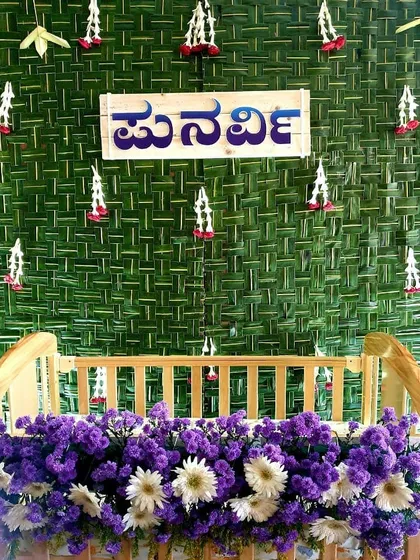 A wooden cradle decorated with purple and white flowers, set against a traditional woven coconut leaf backdrop with a Kannada nameplate.