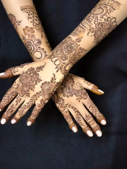 A beautiful pose showcasing two full arms of henna art. This design is perfect for a wedding guest or bridesmaid, with flowing floral vines and leafy patterns.
